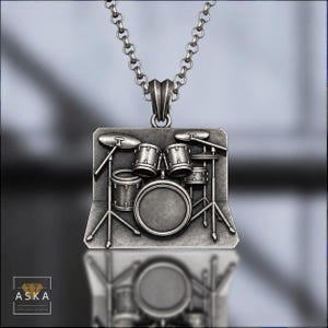 May include: A silver-toned metal pendant necklace featuring a detailed relief of a drum kit. The rectangular pendant hangs from a round link chain. The design depicts a bass drum, snare, toms, and cymbals on stands. The text ASKA is visible.
