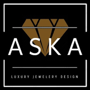 Op de afbeelding: Logo voor luxe sieradenontwerp, met het woord "ASKA" in witte hoofdletters boven de woorden "LUXURY JEWELRY DESIGN" in kleinere witte letters. Het logo bevat een bruine diamantvorm en is geplaatst tegen een zwarte achtergrond, omlijst door een witte rand.