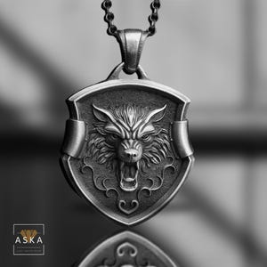 Op de afbeelding: Een zilveren ketting met een hanger met een wolfskop in een schildontwerp. De wolfskop is gedetailleerd en realistisch, met een grommende uitdrukking. Het schild is versierd met ingewikkelde krulwerk.