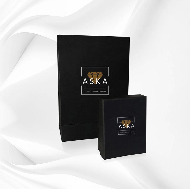Op de afbeelding: Twee zwarte sieradendoosjes van verschillende formaten. Beide dozen hebben het woord "ASKA" in het wit, met een gouden diamantafbeelding en de tekst "LUXURY JEWELLERY DESIGN" in het wit. De dozen staan op een witte achtergrond.