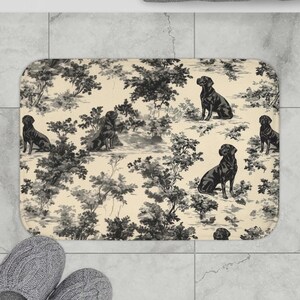Puede incluir: Alfombra de baño rectangular con un diseño toile en blanco y negro. El diseño presenta perros Labrador Retriever negros y árboles sobre un fondo crema. La alfombra está hecha de material suave y absorbente y tiene un respaldo antideslizante.