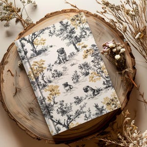 May include: A white journal with a black and gold pattern of pugs and trees. The journal is on a wooden surface.
