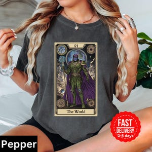 Puede incluir: Una camiseta gris oscuro con un diseño de carta de tarot que presenta una figura morada y verde sosteniendo un bastón. La carta está etiquetada como "21 El Mundo".