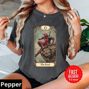 May include: A dark gray t-shirt with a graphic of Deadpool as The Fool tarot card. The card is in a vintage style with a gold frame and the number 0 in the top corner. The text "The Fool" is printed below the image.