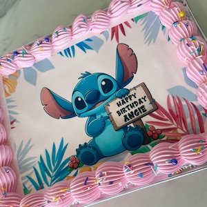 May include: Rectangular cake with pink frosting border and a cartoon image of Stitch holding a sign that reads "Happy Birthday Angie." The cake has a tropical floral design background and is decorated with sprinkles.