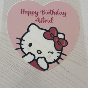 May include: A pink heart-shaped sticker featuring a cartoon image of Hello Kitty with a red bow. The text "Happy Birthday Astrid" is written in a cursive font above the character. The sticker is on a light-colored surface.