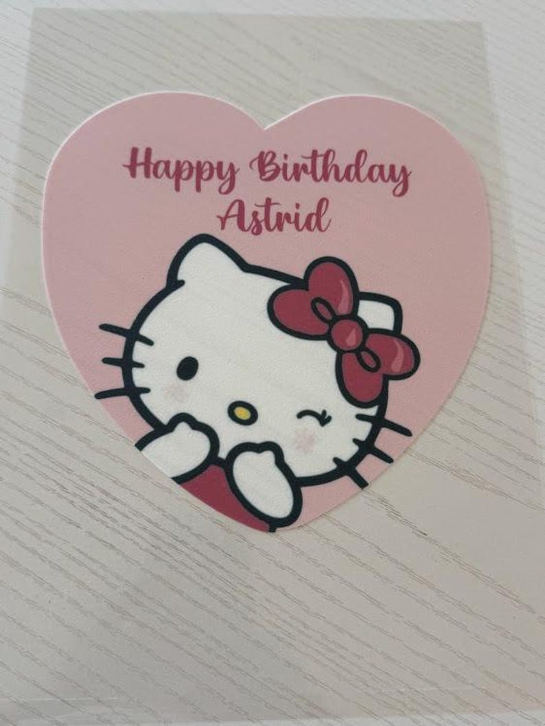 May include: A pink heart-shaped sticker featuring a cartoon image of Hello Kitty with a red bow. The text "Happy Birthday Astrid" is written in a cursive font above the character. The sticker is on a light-colored surface.