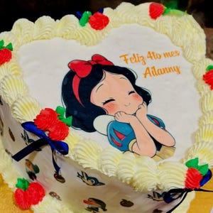 May include: A heart-shaped cake decorated with white frosting and a cartoon image of Snow White. The cake is adorned with red roses and a blue ribbon. The text "Feliz 4to mes Ailanny" is written on the cake.