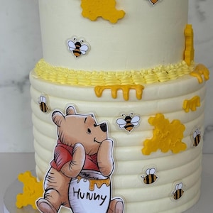 Puede incluir: Un pastel de dos pisos con glaseado blanco y detalles amarillos. El pastel está decorado con abejas de dibujos animados, formas de panal y un personaje de Winnie the Pooh sosteniendo un tarro de miel que dice "Hunny".