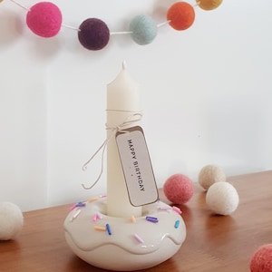Könnte beinhalten: Eine weiße Kerze in einem donut-förmigen Halter mit bunten Streuseln. Die Kerze hat ein Etikett mit der Aufschrift "HAPPY BIRTHDAY". Eine Girlande aus bunten Filzkugeln hängt im Hintergrund. Mehrere Filzkugeln liegen auf der Holzoberfläche.