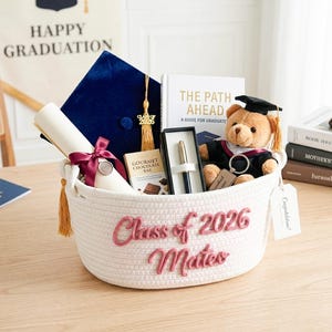 May include: A graduation gift basket with a blue cap, diploma, teddy bear in a graduation gown, pen, and chocolate bar. The white basket has "Class of 2026 Mateo" in pink lettering. A sign reads "Happy Graduation".