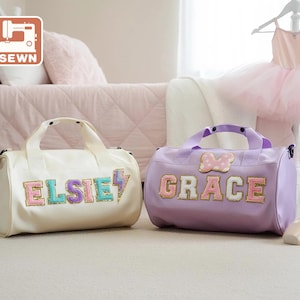 Könnte beinhalten: Zwei personalisierte Sporttaschen, eine cremefarbene und eine lavendelfarbene, stehen nebeneinander. Die cremefarbene Tasche trägt den Namen "ELSIE" mit einem Blitz, und die lavendelfarbene Tasche trägt den Namen "GRACE" mit einer Schleife. Ein Balletttutu und -schuhe sind im Hintergrund.