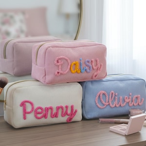 May include: A collection of corduroy cosmetic bags in various pastel colors, including pink, white, and blue. Each bag features a name embroidered in a contrasting color. The bags are arranged on a wooden surface, with makeup brushes and other beauty products nearby.