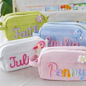 Op de afbeelding: Vier corduroy make-up tasjes in pastelkleuren: groen, blauw, wit en roze. Elke tas heeft een geborduurde naam: Faith, Emilia, Julie en Penny. De tasjes zijn versierd met bloemen en snoepjes.