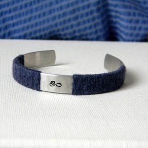 Op de afbeelding: Een zilverkleurige metalen armband met een platte middenplaat gegraveerd met een klein oneindigheidssymbool en een hartje. De zijkanten van de armband zijn omwikkeld met donkerblauw garen, wat een gestructureerd contrast vormt met de gladde metalen afwerking.