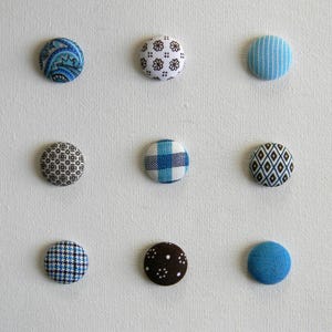 Op de afbeelding: Negen met stof bedekte knopen in verschillende patronen en kleuren, waaronder blauwe paisley, witte bloemen en blauw geruit. De knopen zijn gerangschikt op een witte achtergrond, die hun diverse ontwerpen laat zien.