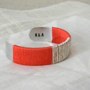 May include: A silver cuff bracelet with a vibrant orange and silver wrapped design. The bracelet has the initials "A & A" stamped on the inside. The bracelet is resting on a white, textured surface.