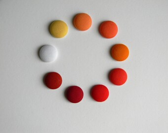 May include: A collection of nine fabric-covered buttons arranged in a circle. The buttons vary in color, including yellow, orange, red, and white. The buttons are approximately 1 inch in diameter and are set against a white background.