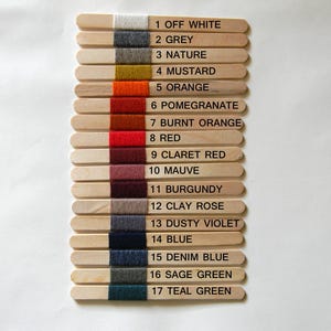 May include: Seventeen wooden craft sticks arranged vertically, each wrapped with a different colored yarn sample. The sticks are numbered 1 through 17 with corresponding color names printed in black text next to each yarn swatch, ranging from off white to teal green.