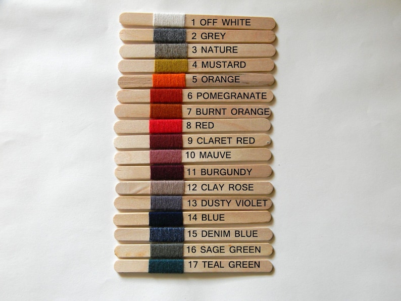 May include: Seventeen wooden craft sticks arranged vertically, each wrapped with a different colored yarn sample. The sticks are numbered 1 through 17 with corresponding color names printed in black text next to each yarn swatch, ranging from off white to teal green.