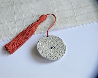 May include: A circular silver-toned metal charm with a hammered, textured surface featuring the letters CG stamped in the center. A thin cord connects the charm to a long, rust-colored fabric tassel that hangs beside the metal disc.