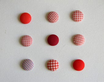 May include: A set of nine circular fabric-covered buttons arranged in a three-by-three grid. The buttons feature various red and white patterns, including solid red, small geometric shapes, a red and white gingham check, and a blue and red polka dot.