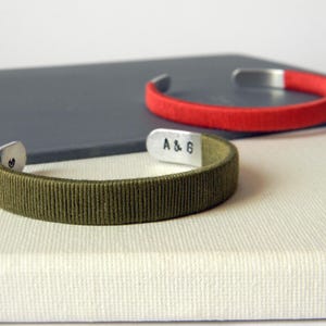 Puede incluir: Dos pulseras de puño. Una es verde oliva con una banda de tela texturizada y extremos plateados. La otra es roja con una banda lisa y extremos plateados. La pulsera verde oliva tiene las letras "A & 6" estampadas.