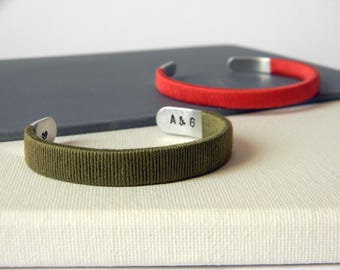 May include: Two cuff bracelets featuring metal ends and wrapped cord bands. One bracelet is olive green with a heart symbol stamped on the metal, and the other is bright red. The olive bracelet has the text A & G stamped on the inner metal end.
