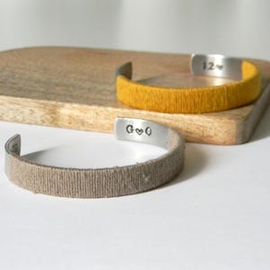 May include: Two open cuff bracelets with textured bands. One is mustard yellow, the other is tan. The metal cuffs have stamped letters and numbers. The bracelets rest on a wooden chopping board.