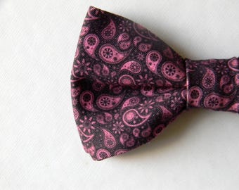 May include: A bow tie featuring a dark black base color with a repeating pattern of pink paisley shapes and small floral accents. The fabric has a visible woven texture, and the bow tie is tied in a classic shape with a central knot detail.