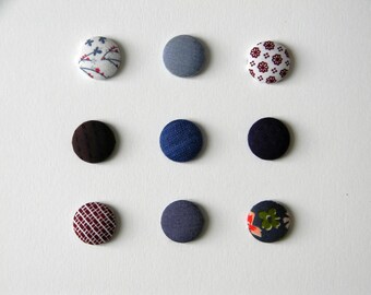 May include: Nine fabric-covered buttons in various colors and patterns are arranged on a white surface. The buttons feature floral, geometric, and solid designs in shades of blue, brown, and red. These buttons are ideal for sewing and craft projects.