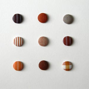 Op de afbeelding: Negen met stof bedekte knopen in verschillende kleuren en patronen. De knopen bevatten strepen, effen kleuren en stoffen met textuur in tinten bruin, oranje en beige. De knopen zijn gerangschikt in een raster van drie bij drie op een witte achtergrond.