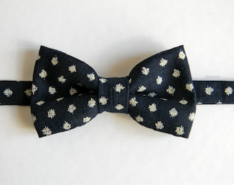 May include: A dark navy blue bow tie featuring a repeating pattern of small, off-white, cross-shaped embroidered dots. The bow tie has a textured fabric appearance and includes a matching horizontal strap extending from both sides of the center knot.