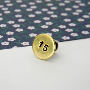 Puede incluir: Un pin redondo de color latón con el número "15" en negro. El pin está sobre una superficie blanca con un fondo estampado floral en azul marino y blanco.