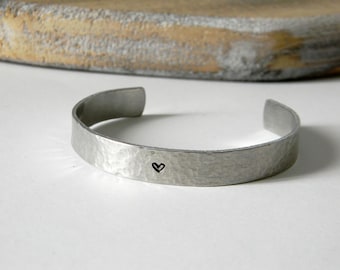 May include: A silver-toned metal cuff bracelet with a hammered, textured finish. The bracelet features a small, simple black heart symbol stamped onto the center of the outer surface. The band is open-ended, forming a C-shape design.