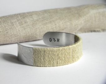 May include: A silver-toned metal cuff bracelet featuring a smooth, polished finish on the ends and a central section wrapped in light beige twine. The interior of the metal band is stamped with the black uppercase letters D & N.