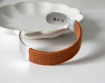 May include: A silver-toned metal cuff bracelet featuring a smooth, polished finish on the ends and a textured, rust-colored thread wrapped around the center. The interior of one end is stamped with the letters E and G separated by a heart symbol.