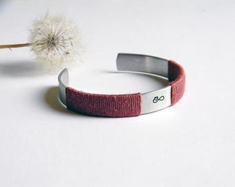 May include: A silver-toned metal cuff bracelet featuring a section wrapped in dark red thread. The metal surface displays a small, black engraved infinity symbol with a tiny heart integrated into the loop design, positioned near the thread wrap.