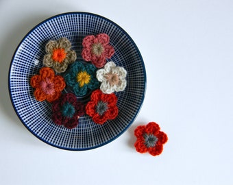 May include: A collection of handmade crochet flowers in various colors, including orange, red, teal, and beige, arranged on a blue and white patterned plate. One flower is separate from the others. The flowers have a detailed, textured appearance.
