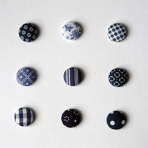 May include: Nine fabric-covered buttons in various blue and white patterns. Designs include polka dots, floral, geometric, and striped patterns. The buttons are arranged on a white surface, showcasing their diverse designs and textures.