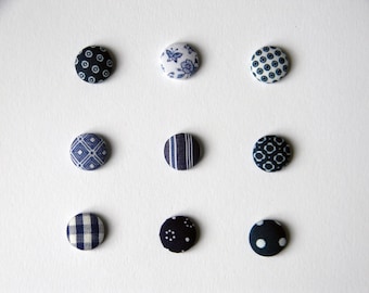 May include: A set of nine circular fabric-covered buttons arranged in a three-by-three grid. Each button features a distinct navy blue and white pattern, including floral motifs, geometric shapes, stripes, gingham checks, and polka dots.