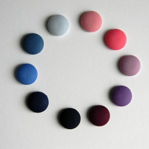 Könnte beinhalten: Eine Anordnung von zehn stoffbezogenen Knöpfen in einem Kreis. Die Farben variieren von Blau über Rosa und Lila bis Schwarz. Die Knöpfe sind rund und haben eine strukturierte Oberfläche.