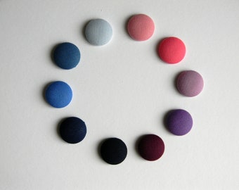 May include: A collection of ten fabric-covered buttons in a variety of colors, arranged in a circular pattern. The colors include shades of blue, pink, purple, and black. The buttons are round and have a textured appearance.