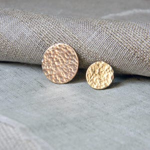 Könnte beinhalten: Zwei goldfarbene, gehämmerten Metall-Ohrstecker. Einer ist größer und rund, der andere kleiner. Die Ohrringe haben eine strukturierte Oberfläche und werden auf einem neutralfarbenen, strukturierten Stoffhintergrund präsentiert.