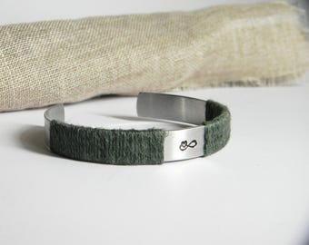 May include: A silver-toned metal cuff bracelet featuring two sections wrapped in dark olive green yarn. The metal surface displays a small, stamped black infinity symbol with a heart shape integrated into the loop on the front side.