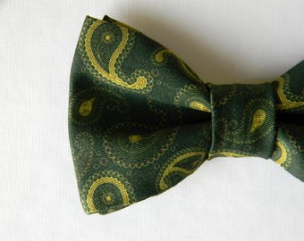 May include: A dark forest green bow tie featuring a repeating golden yellow paisley pattern. The fabric has a visible diagonal weave texture throughout the entire surface of the bow tie, which is tied in a classic shape with a central knot.