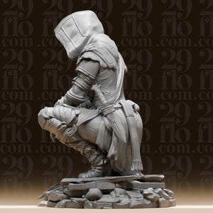 May include: A detailed, grey figurine of a hooded figure in a crouching position, possibly a character from a fantasy or historical setting. The figure is wearing armour and a hood, with a sword at the waist. The base is a textured, rocky surface.