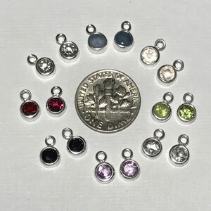 May include: A collection of 14 small silver charms with different colored gemstones. The charms are arranged in a circle around a US dime.