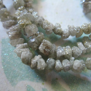 Puede incluir: Una hebra de cuentas de diamante gris en bruto. Las cuentas son irregulares en forma y tama&ntilde;o, y tienen un aspecto natural y sin pulir.