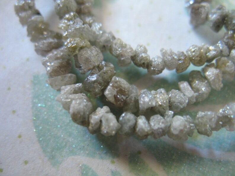Puede incluir: Una hebra de cuentas de diamante gris en bruto. Las cuentas son irregulares en forma y tama&ntilde;o, y tienen un aspecto natural y sin pulir.